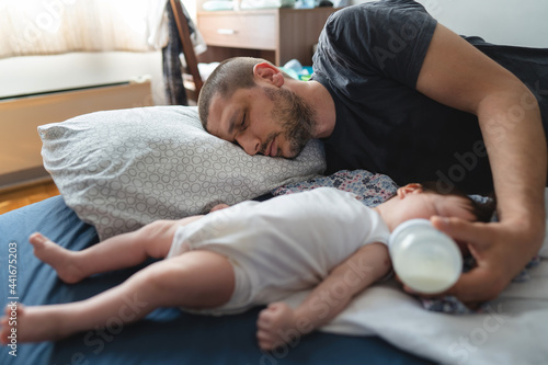 Exhausted man father lying by his three months old baby on the bed at home sleeping while holding baby bottle feeding infant child parenthood concept selective focus