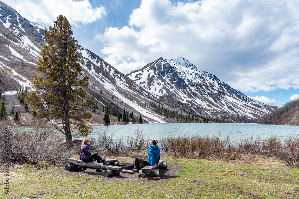 Hikers resting, sitting beside a isolated St Elias Lake in Kluane National Park during spring time, May with no other people. Snow capped mountains surrounding the scenic, Canadian view. 