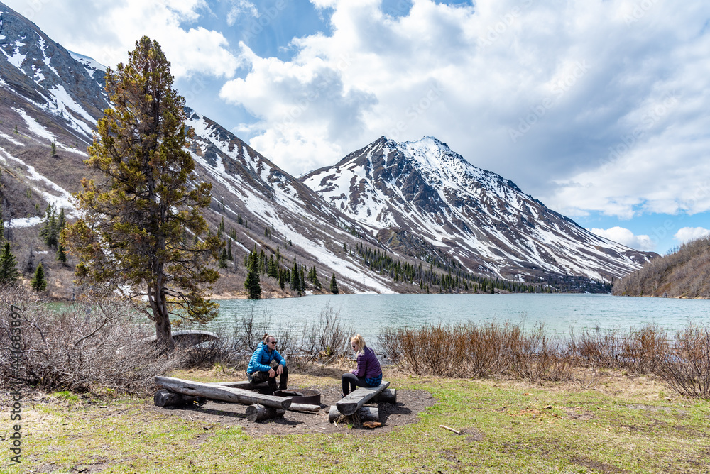 Fototapeta premium Hikers resting, sitting beside a isolated St Elias Lake in Kluane National Park during spring time, May with no other people. Snow capped mountains surrounding the scenic, Canadian view. 