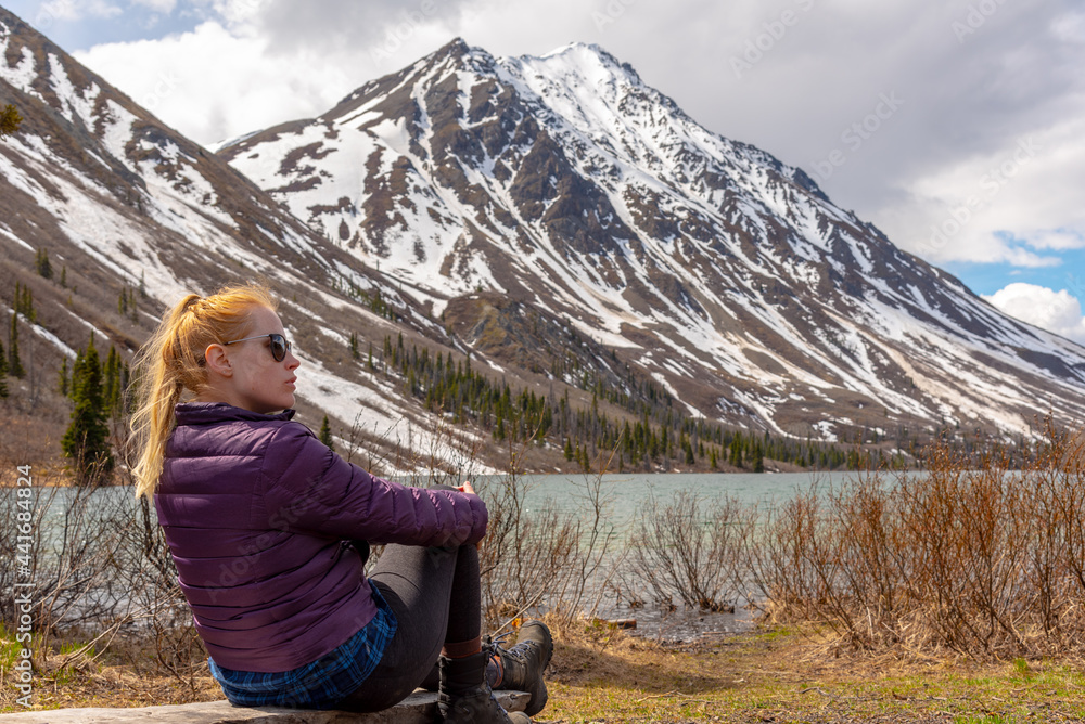 Woman in purple jackets sitting beside a lake with snow capped mountains in background during spring time, May. Wilderness of Yukon Territory in northern Canada.  