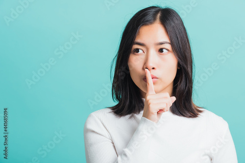 Young beautiful Asian woman holding index finger on her mouth lips, Portrait female hush silence, studio shot isolated on a blue background, Gesture of shhh photo