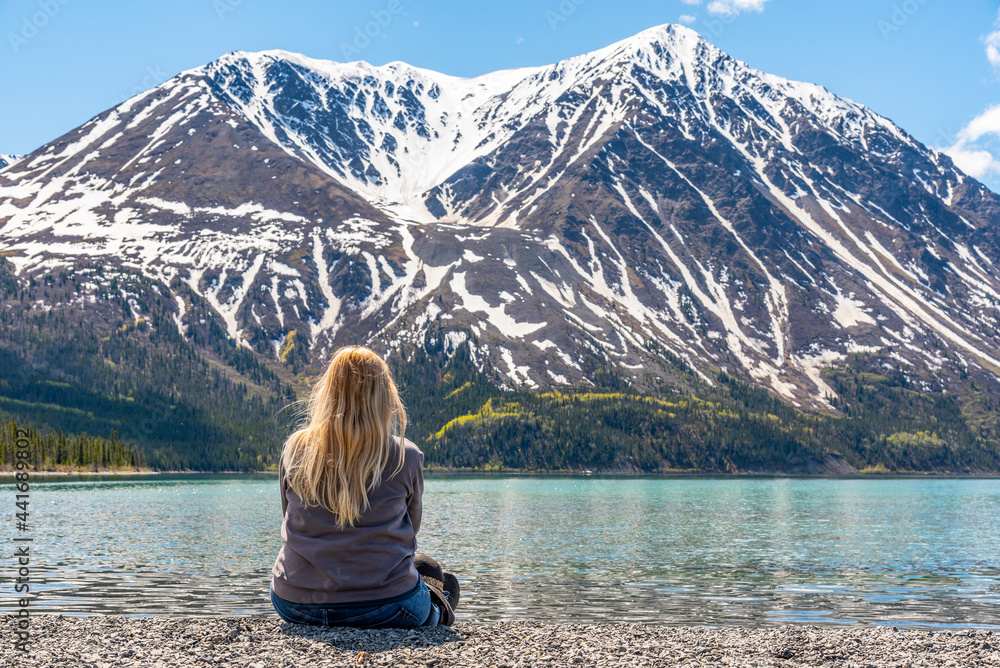Woman with long blonde hair sitting beside a calm, pristine Kathleen Lake in northern Canada during spring time with massive snow capped mountain peaks in scenic background. 