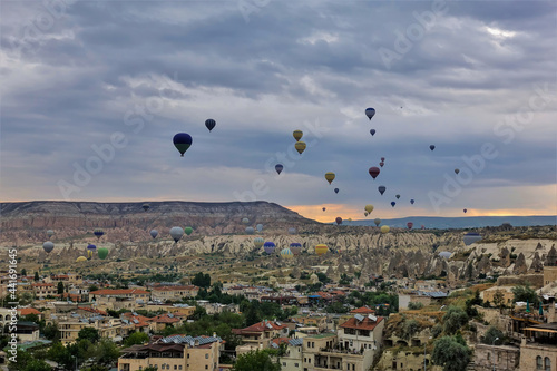 Flying over Cappadocia at dawn. In the cloudy sky, over the village, colorful balloons. The horizon is illuminated in orange. In the distance is a picturesque mountain with a flat top. Turkey