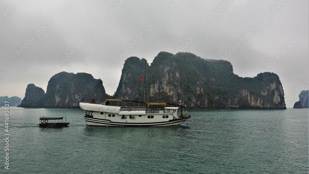 A tourist ship sails on calm water. Quaint islands with sheer rocky slopes hide in the fog. Halong Bay. Vietnam