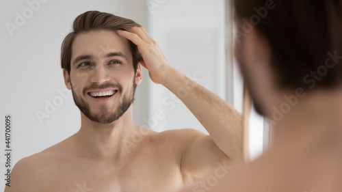 Happy handsome young guy combing smooth brown hair with fingers at mirror in bathroom, looking at reflection with toothy smile. Man satisfied with haircare cosmetic product, enjoying bath routine