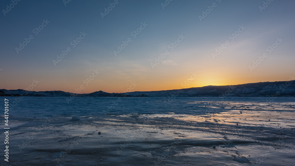 Sunset over a frozen lake. There is snow on the ice and the reflections of the setting sun. The sky above the ridge is highlighted in orange. Baikal