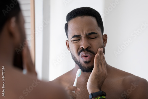 Fotografie Stressed upset mixed raced Black man feeling pain and discomfort while brushing teeth in bathroom, holding toothbrush, touching cheek with painful grimace