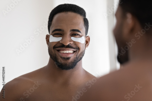 Happy attractive African American guy with applied collagen gel patches on under eye skin smiling at camera. Young man caring for under-eye skin, preventing bags, reducing wrinkles. Head shot portrait photo