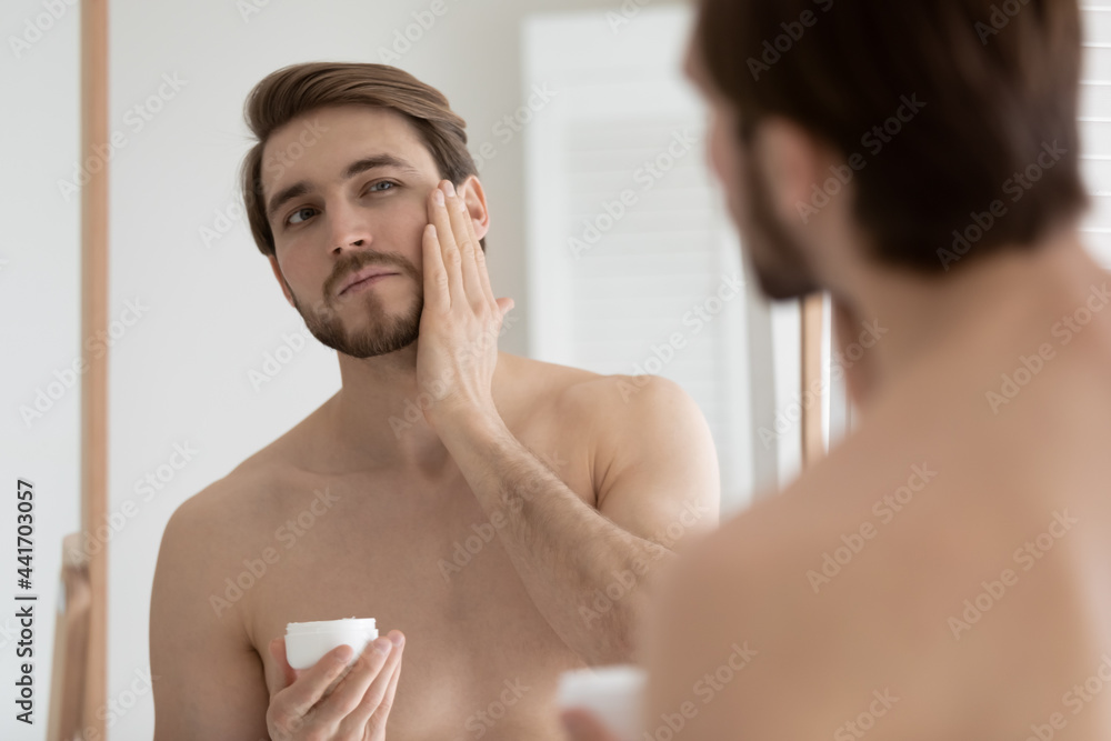 Confident handsome young man with muscular torso applying aftershave balm, lotion, moisturizing night cream, sunscreen, touching face, stubble, holding cosmetic jar, looking in mirror. Confident handsome young man with muscular torso applying aftershave balm, lotion, moisturizing night cream, sunscreen, touching face, stubble, holding cosmetic jar, looking in mirror.