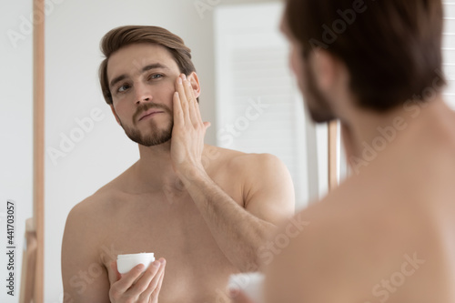 Cuadro en lienzo Confident handsome young man with muscular torso applying aftershave balm, lotion, moisturizing night cream, sunscreen, touching face, stubble, holding cosmetic jar, looking in mirror