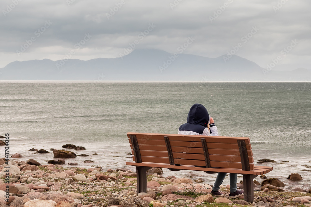 Teenager girl sitting on a bench and enjoys view on Atlantic ocean, low cloudy sky. Croagh Patrick in the background. Travel and tourism concept Teenager girl sitting on a bench and enjoys view on Atlantic ocean, low cloudy sky. Croagh Patrick in the background. Travel and tourism concept