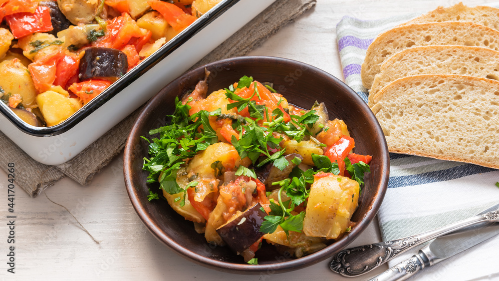Homemade, rustic vegetarian dish is baked potatoes with eggplant and tomatoes on a plate next to a baking dish. Homemade, rustic vegetarian dish is baked potatoes with eggplant and tomatoes on a plate next to a baking dish.