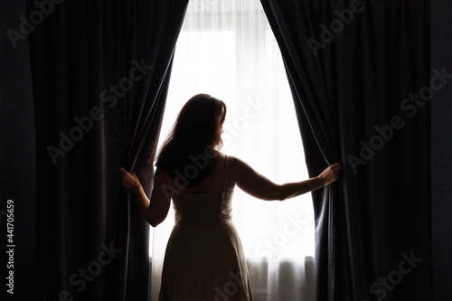 A white woman is opening dark curtains and looking out of window