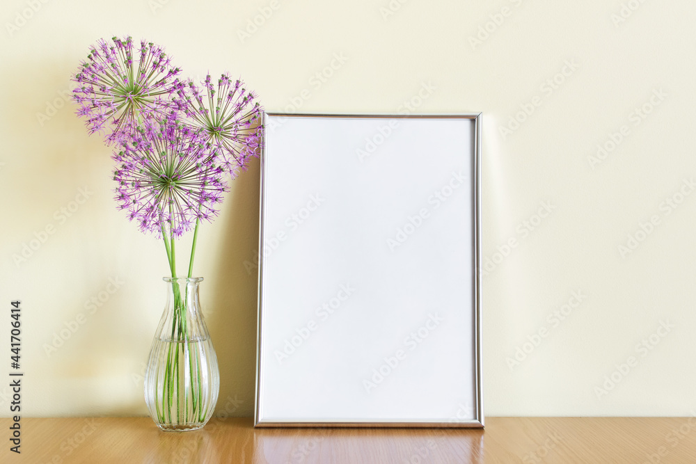 Mockup template with A4 frame standing on wooden shelf with three purple summer flowers branches in glass vase. Mockup template with A4 frame standing on wooden shelf with three purple summer flowers branches in glass vase.
