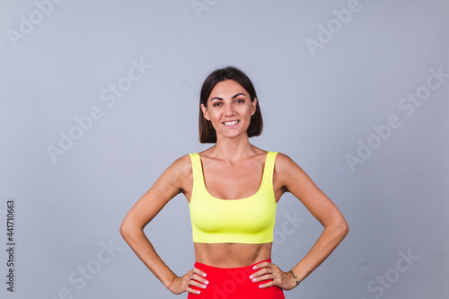 Sports woman stands on gray background, demonstrating her abs, satisfied with the results of fitness training and diet, has a happy facial expression, wears a sports top and tight leggings