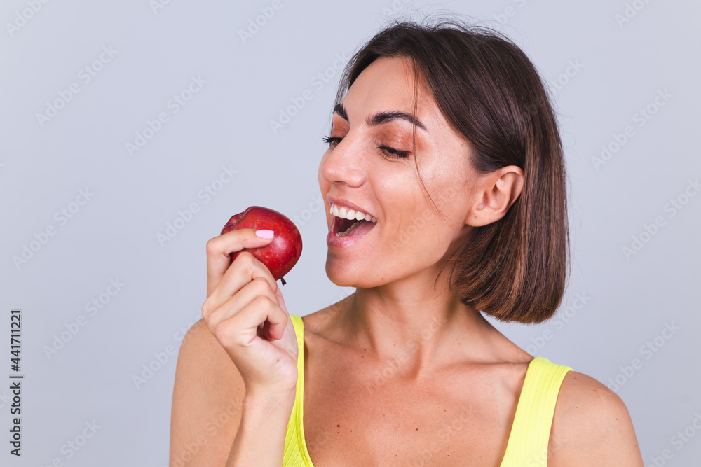Sports woman stands on gray background, satisfied with the results of fitness training and diet, holding scale, wears top and leggings, holds apple Sports woman stands on gray background, satisfied with the results of fitness training and diet, holding scale, wears top and leggings, holds apple