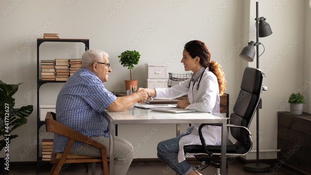 Happy young Caucasian female doctor in white medical uniform handshake old male patient close health insurance deal at meeting. Smiling woman GP shake hand get acquainted with mature man in clinic.