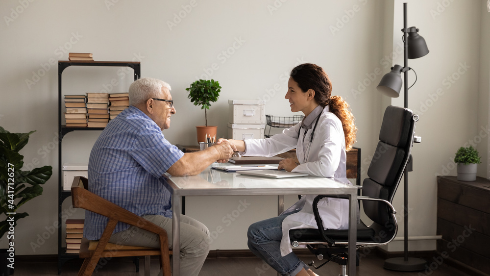 Happy young Caucasian female doctor in white medical uniform handshake old male patient close health insurance deal at meeting. Smiling woman GP shake hand get acquainted with mature man in clinic. Happy young Caucasian female doctor in white medical uniform handshake old male patient close health insurance deal at meeting. Smiling woman GP shake hand get acquainted with mature man in clinic.