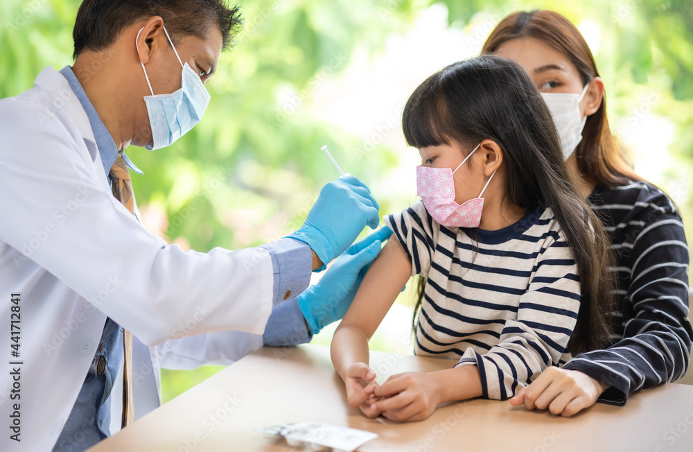 Asian  senior doctor wearing gloves and isolation mask is making a COVID-19 vaccination in the shoulder of child patient with her mother at hospital.