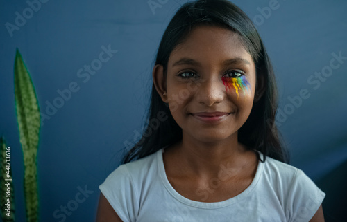 Portrait of a cheerful young member of LGBTQ+ community smiling with pride flag drawn under the eyes| proud, confident teenage girl in a positive mood in front of a solid background