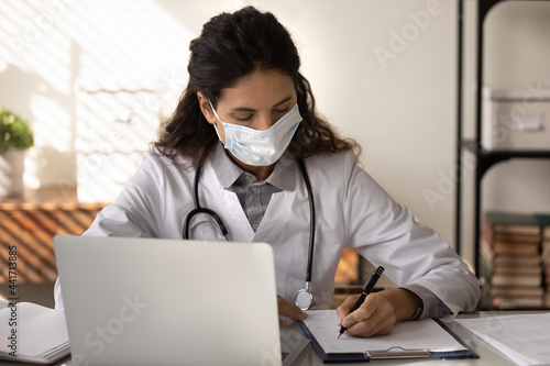 Young Caucasian female doctor in medical facemask work in clinic on computer write in journal. Woman nurse or GP in facial mask against corona virus busy in hospital. Covid-19, corona concept.