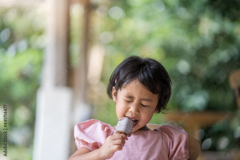Adorable little Asian girl sitting on wooden chair eating chocolate ice pop Adorable little Asian girl sitting on wooden chair eating chocolate ice pop
