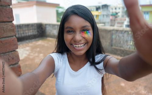 A playful dark skinned young girl of LGBTQ community with rainbow eye makeup laughing and taking a selfie | teen girl smiling cheerfully and giving hugs