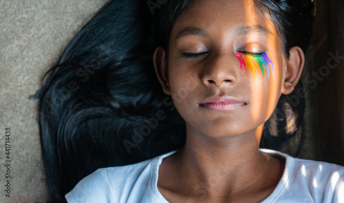 Close-up shot of a young dark skinned girl with sunlight falling on her face with rainbow makeup on closed eyes | proud LGBTQ community, gender fluidity, pride concept