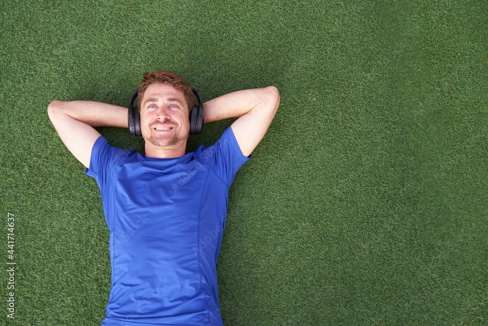 Close up portrait of smiling man lying on grass while listening to music an wearing sunglasses.Happy man lying with both hands resting behind his neck on the grass Close up portrait of smiling man lying on grass while listening to music an wearing sunglasses.Happy man lying with both hands resting behind his neck on the grass