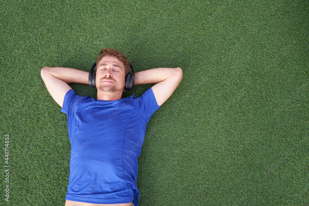 Close up portrait of smiling man lying on grass while listening to music an wearing sunglasses.Happy man lying with both hands resting behind his neck on the grass Close up portrait of smiling man lying on grass while listening to music an wearing sunglasses.Happy man lying with both hands resting behind his neck on the grass
