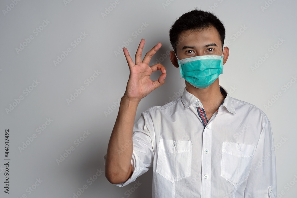 Portrait of young asian man wearing health mask showing OK gesture in front of camera against white background. Agreement gesture in healthcare concept. Novel Coronavirus (COVID-19) Portrait of young asian man wearing health mask showing OK gesture in front of camera against white background. Agreement gesture in healthcare concept. Novel Coronavirus (COVID-19)