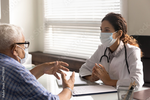 Young Caucasian female doctor in facial mask and old male patient talk discuss disease anamnesis in hospital. Woman GP and mature man in facemasks have consultation in clinic in covid-19 corona times.