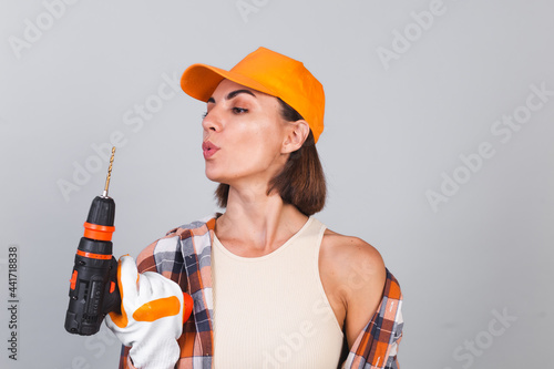 Beautiful woman in plaid shirt, hat and gloves, on gray background holds drill to repair  home confident happy smile © Анастасия Каргаполов