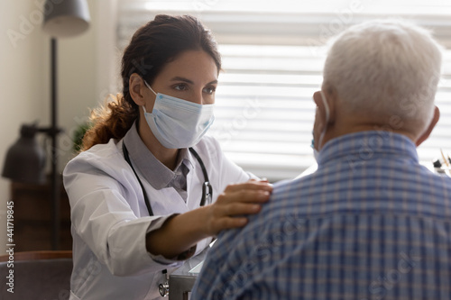 Caring female GP in facial mask touch support old male patient hear bad negative diagnosis at consultation. Woman doctor in facemask comfort caress mature man in hospital  show good medical service.