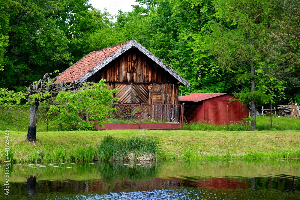 A view of a small wooden shack, hut or house made entirely out of logs and planks with an angled roof and a small storage unit nearby located next to the bank of a vast yet shallow river in Poland A view of a small wooden shack, hut or house made entirely out of logs and planks with an angled roof and a small storage unit nearby located next to the bank of a vast yet shallow river in Poland