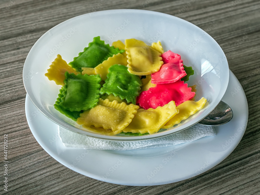 White plate of multi-colored ravioli is standing on a wooden table. Close-up. White plate of multi-colored ravioli is standing on a wooden table. Close-up.