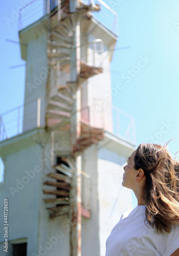 young woman looking at old lighthouse with stairs. alone travelling. local travel. vertical size. side view