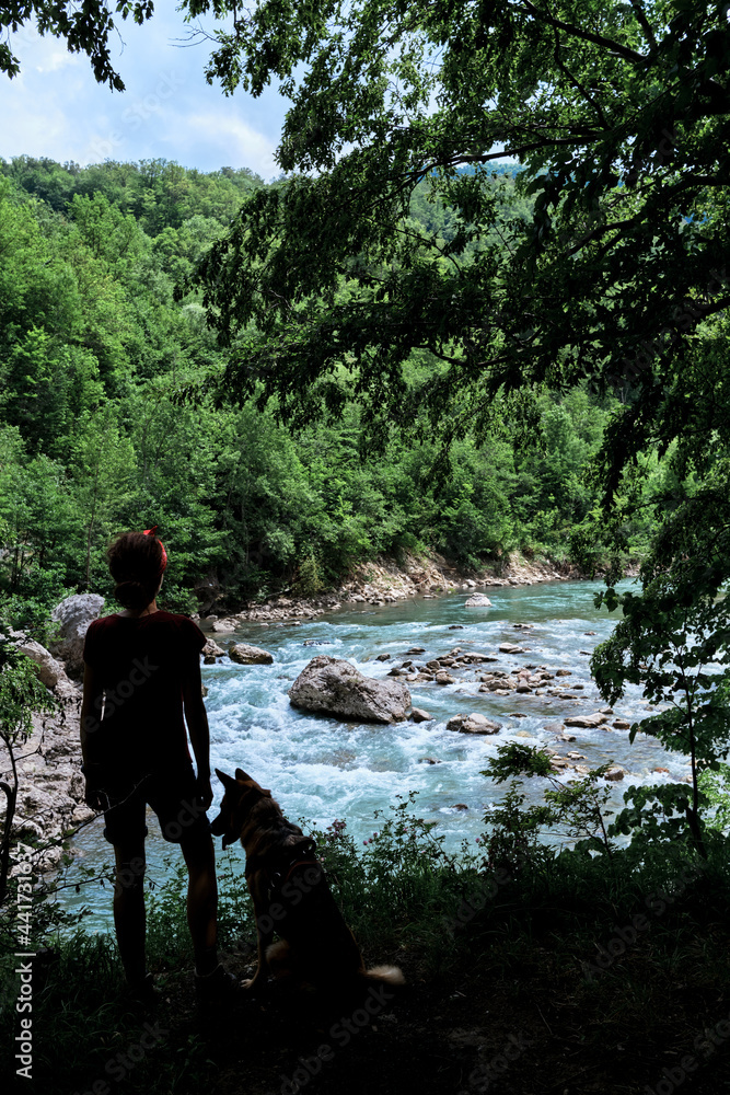 Obraz premium Viewing platform on mountain and female tourist. Traveler girl and German Shepherd dog stand together on cliff and look at stormy beautiful mountain river flowing at bottom of stone gorge.