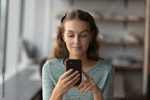 Attractive young woman looking at smartphone screen, reading media news, shopping in internet store, communicating distantly in social network, using mobile software applications or web surfing.