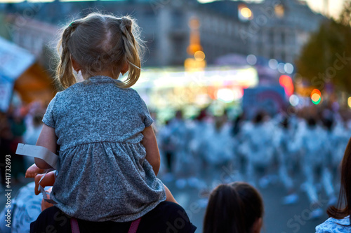 A girl on the shoulders of a parent at a city party. A little blonde girl looks at the concert from the back. A child at a show in the evening on the street. City day and open air.