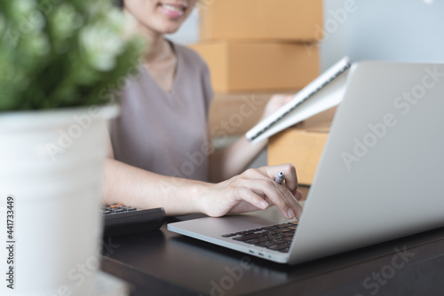 E-commerce, online business concept. Smiling young asian woman entrepreneur checkimng order on laptop and preparing product parcels to deliver to customer