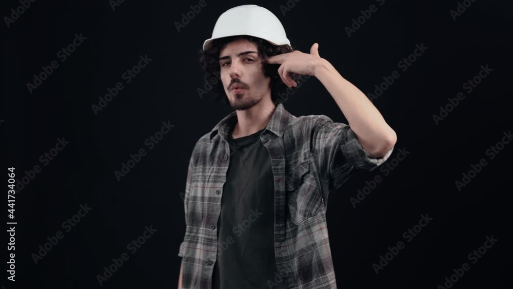 The charismatic young man in the white helmet of an engineer takes his fingers to the temple in the form of a pistol. Isolated on black background. The concept of life. People's emotions. 4k portrait The charismatic young man in the white helmet of an engineer takes his fingers to the temple in the form of a pistol. Isolated on black background. The concept of life. People's emotions. 4k portrait