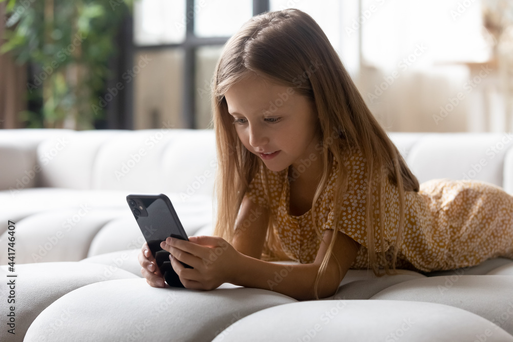 Addicted to modern tech preschool adorable small kid girl lying on cozy couch, involved in playing online mobile games alone at home. Joyful little preschool child having fun online in living room. Addicted to modern tech preschool adorable small kid girl lying on cozy couch, involved in playing online mobile games alone at home. Joyful little preschool child having fun online in living room.
