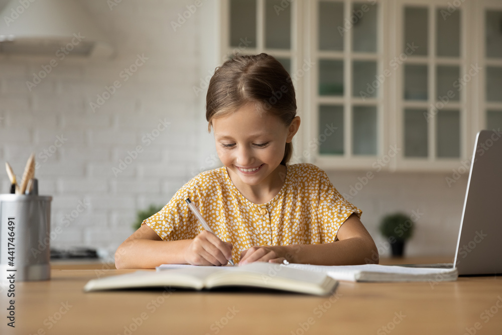Happy cute small 7s child girl doing homework, studying distantly in online school using computer application, handwriting notes in copybook or preparing for exams alone in modern kitchen room. Happy cute small 7s child girl doing homework, studying distantly in online school using computer application, handwriting notes in copybook or preparing for exams alone in modern kitchen room.