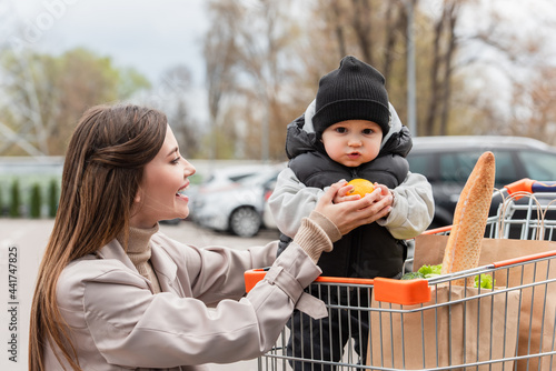 happy woman looking at baby boy holding fresh orange while sitting in shopping cart © LIGHTFIELD STUDIOS