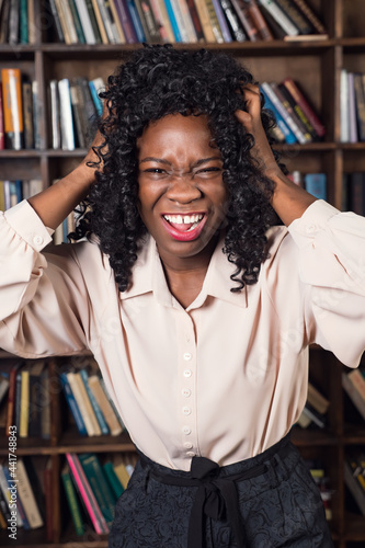Young african american woman holding head in horror against the background of shelves with books.