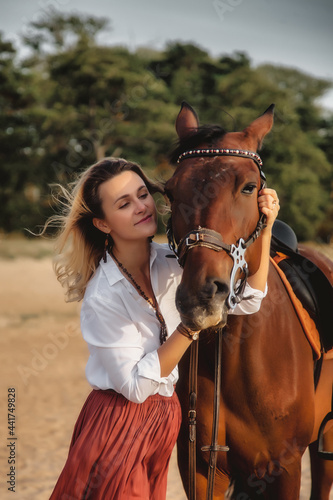 Cute happy young woman with horse in summer beach by sea. Rider female drives her horseback in nature on evening sunset light background. Concept of outdoor riding, sports and recreation. Copy space
