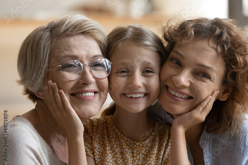 Head shot close up portrait of sincere emotional happy three female generations bonding family enjoying tender sweet moment at home. Joyful candid little kid girl cuddling smiling granny and mommy.