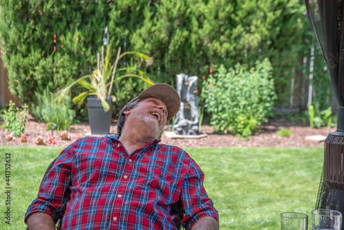 A bearded, handsome gay man in his 50s throws his head back in laughter as he enjoys an afternoon relaxing on his back yard patio. Room for copy.