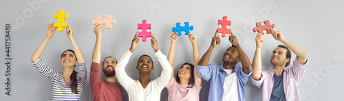 Group of six happy smiling young people holding big colorful jigsaw puzzle pieces standing against light grey studio background. Team of creative millennials cooperate and find good solution together
